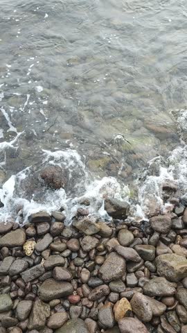 Top view of colorful pebbles under clear shallow water with gentle sea waves washing ashore. Natural rocky beach scenery.