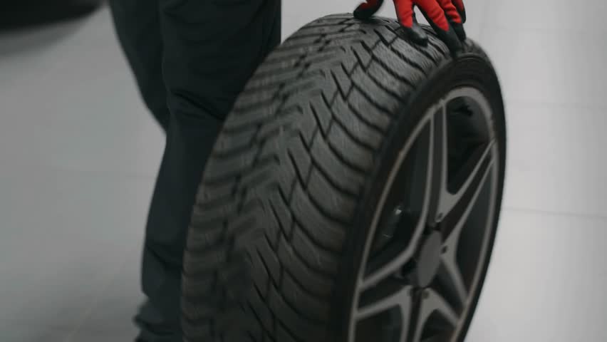 Medium shot of legs and hands of a male mechanic in gray overalls and red gloves rolling a car tire, highlighting automotive work, precision, and hands-on labor.