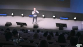 Blurred view of speaker presenting on stage at conference in auditorium, audience seated, people work on laptops, stage lights glow, large screen behind speaker - Powered by Shutterstock - Get 15% off with code: PIKWIZARD15