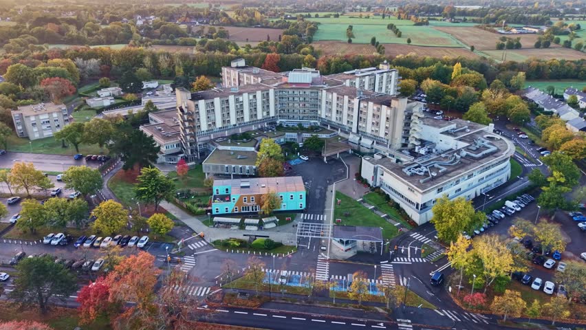 Drone establishing shot of CHU Hôpital Sud in Rennes, France, showing the sprawling hospital complex surrounded by tree-lined roads, parking areas, and green countryside on a sunny autumn morning.