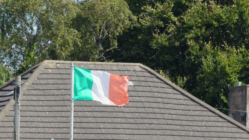 Irish tricolour flag flutters on rooftop flagpole, sunny summer day, residential Edinburgh, steady shot