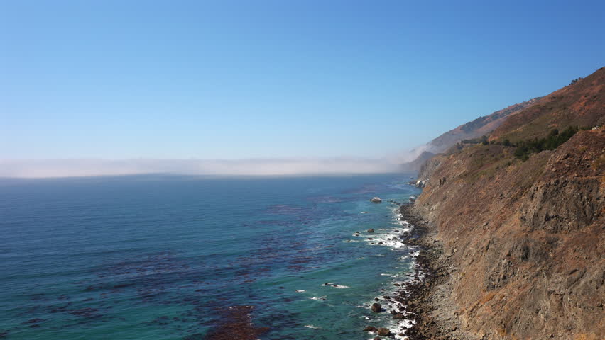 Fog slowly rolls over the steep Big Sur California cliffs as waves hit the rocky shoreline creating a calm coastal timelapse scene captured in soft daylight with cinematic aerial motion