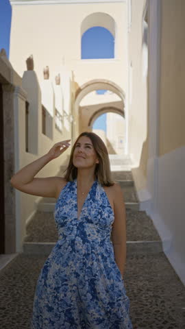 Woman in blue floral dress smiling and looking up on santorini street with arched white buildings; travel serenity.