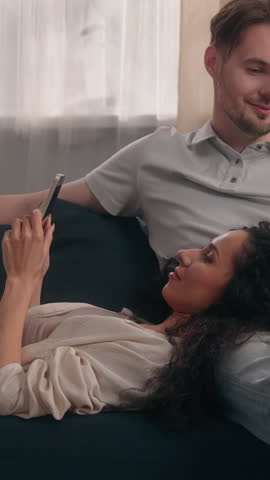 Multiracial couple at home with Caucasian man using laptop addicted to internet while Hispanic woman on couch chatting on mobile phone smiling laughing social media addiction family gadget indoor room