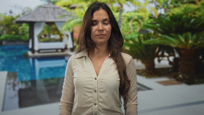 Woman standing by pool at hotel building outdoors, wearing beige button shirt, facing camera with neutral expression; serenity.