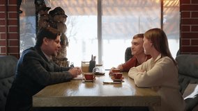 Young people at wooden table in modern cafe with large windows, woman in beige sweater gestures actively, man in jacket smiling, red coffee cups, winter tree view, cozy atmosphere, social gathering. - Powered by Shutterstock - Get 15% off with code: PIKWIZARD15