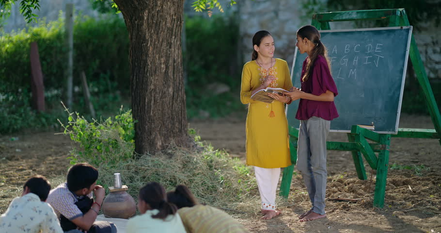 Indian young teen female standing talk do study hold book help to read learn under tree outside rural area. Student girl boy group sitting on mat enjoy break time have fun joy open class air filed