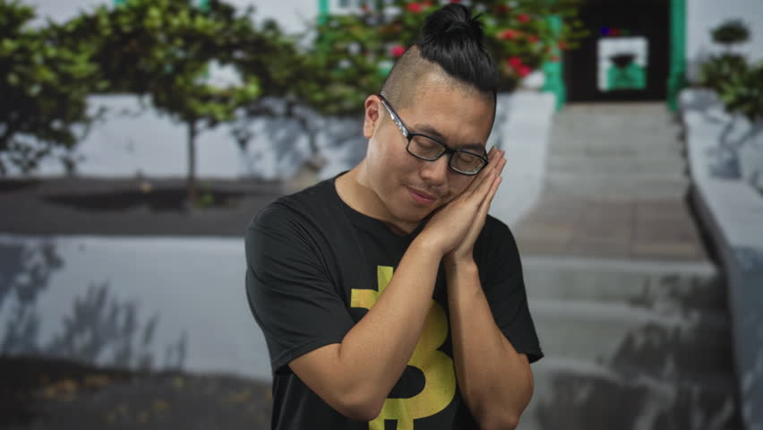 Man in bitcoin t shirt wearing glasses and resting head on hands while standing on building steps; sleepy crypto enthusiast.