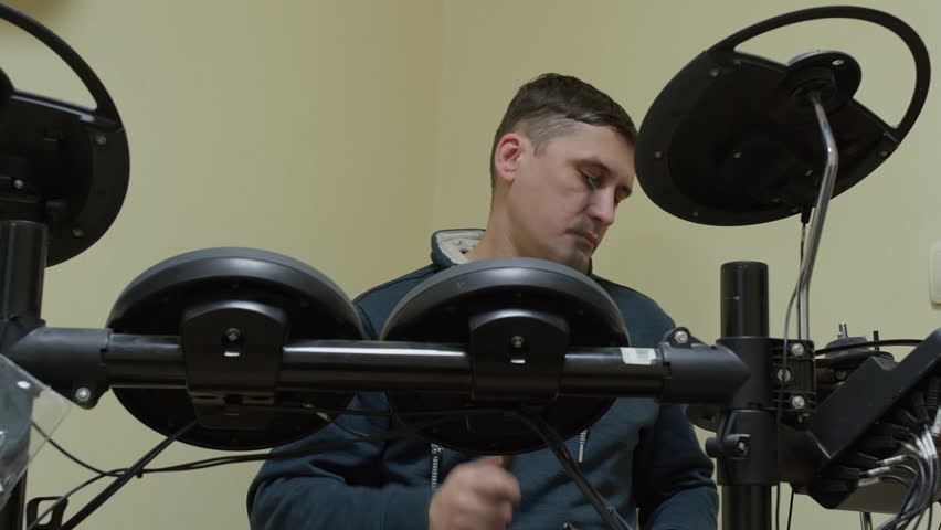 Young male musician in dark blue hoodie sitting at desk with black electronic drum pads and wheels, concentrated expression, neutral beige wall, studio lamps visible, home music production setup.