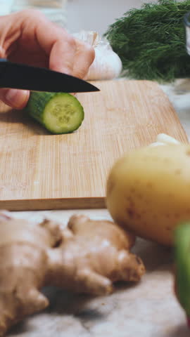 vertical shot. A chef cuts a spring salad of cucumbers and herbs. A man cuts cucumbers for the salad on a wooden cutting board on a kitchen table surrounded by vegetables. A man in the kitchen.