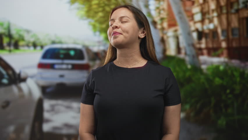 Woman smiling with eyes closed by a parked car on a city street in a black t shirt; serenity mindfulness.
