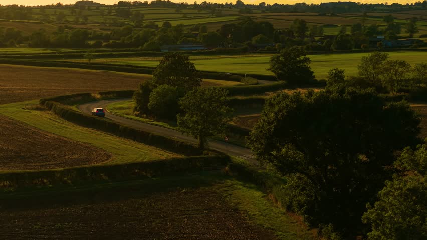 Blue car curving along countryside road in golden sunset fields. Road driving, car traveling, countryside lifestyle vacation. Compact vehicle navigating rural winding roadway during evening glow