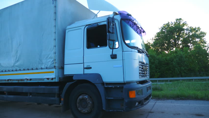 Truck with cargo trailer driving on highway and transporting goods. White lorry riding to destination through countryside at sunset time. Bright sunset light shining at background. Side view Slow mo