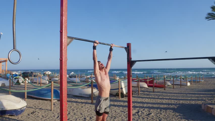 Young shirtless athlete performing pull ups on a horizontal bar. Man with a strong body training outdoors with the sea in the background