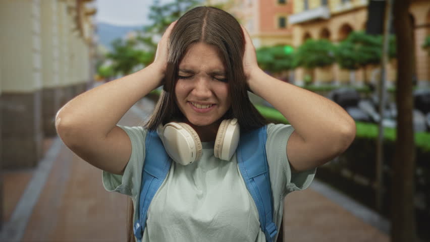 Caucasian teenage girl wearing blue backpack and white headphones covers ears with hands on sunlit city street; stress.