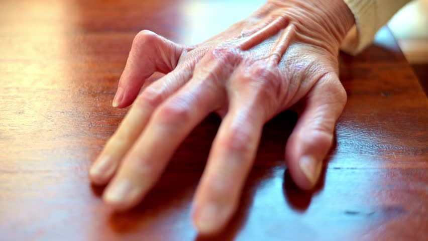 Close-up of an elderly hand resting on a wooden table with deep wrinkles and texture, and a trigger finger.