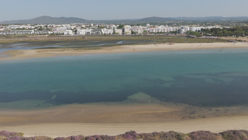 Drone moves forward above shallow water toward Fuzeta beach, passing sandbanks and swimmers before reaching the coastal buildings.