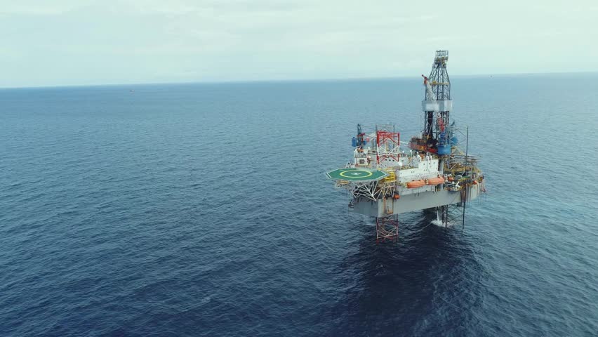 Aerial view captures oil rig amidst open ocean. Structure showcases advanced equipment and operational readiness. Calm waters reflect overcast sky, creating serene backdrop for energy production.