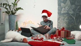 Happy child in Santa hat enjoying Christmas atmosphere at home, sitting with notebook and presents on cozy bed surrounded by warm lights and decorations - Powered by Shutterstock - Get 15% off with code: PIKWIZARD15