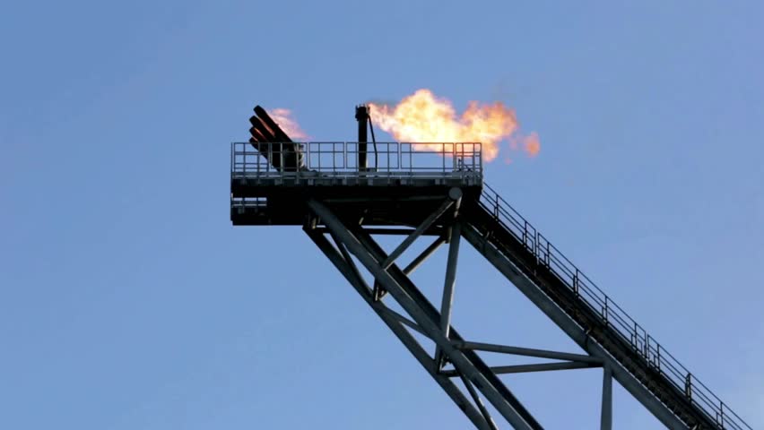 Flames erupt from flare stack atop oil rig creating striking visuals against serene sky. Industrial activity showcased in natural light adds dramatic effect to expansive offshore landscape.