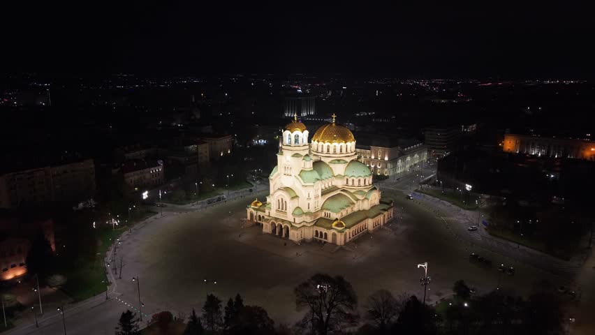 Drone aerial view approaching the illuminated Alexander Nevsky Cathedral from above, showcasing its golden domes and the glowing night cityscape of Sofia.