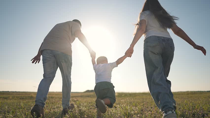 Dog jumps near boy while father holds toddler hand. Kid moves ahead. Family follows dog. Son runs to mother. Parent guides child. Dog circles group. Happy family walks through bright summer field.