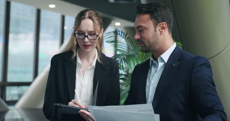Two happy corporate colleagues laugh joyfully while reviewing business papers and charts together in a modern office building demonstrating successful teamwork and positive work atmosphere
