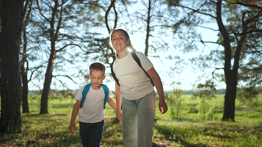Girl leads boy child walk through forest. Backpack on sister, boy happy smile under trees. Forest hike together brother sister. Nature sunlight on walk. Child boy sister laugh in trees forest.