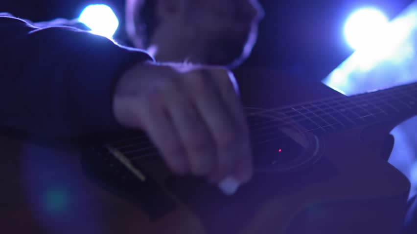 A musician plays an acoustic guitar on stage with spotlights in the background