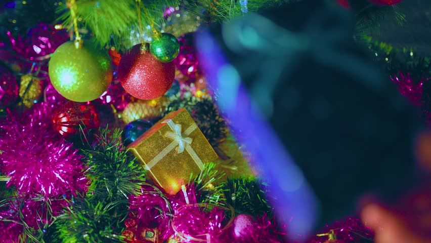 An unrecognizable hand of a person puts a box with a gift next to another under the tree. Red, green and yellow toys hang on the branches. Holidays coming soon. selective focus.
