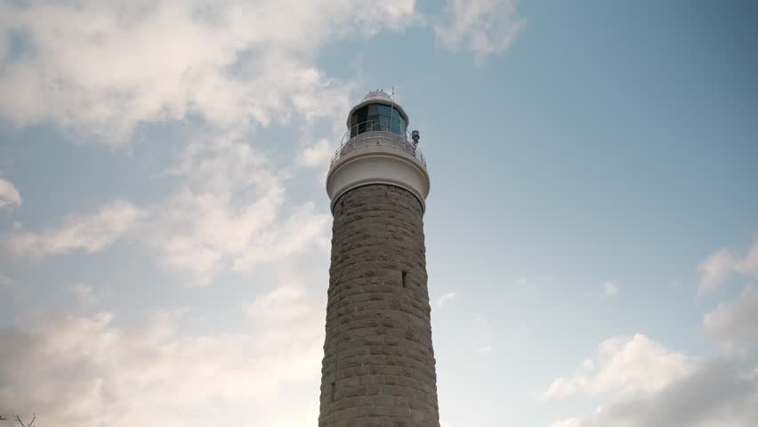Low angle view looking up at a historic stone lighthouse tower against a blue sky with moving clouds. Majestic maritime architecture and coastal navigation beacon standing tall.