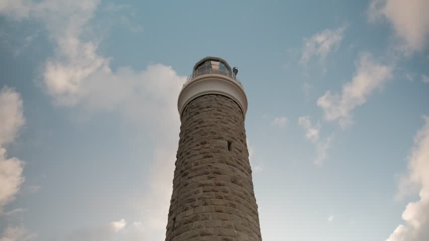Low angle view looking up at a historic stone lighthouse tower against a blue sky with moving clouds. Majestic maritime architecture and coastal navigation beacon standing tall.