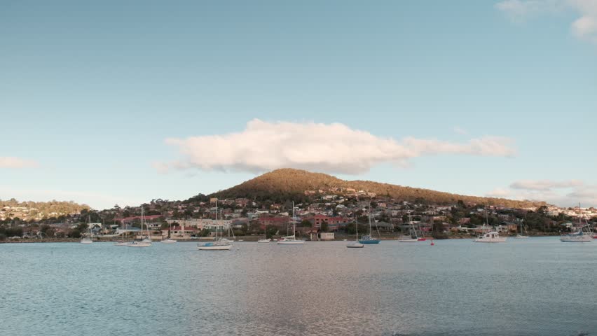 Scenic view of sailboats moored on the Derwent River in Hobart, Tasmania. Warm sunlight illuminates a residential hillside topped with a single large white cloud.