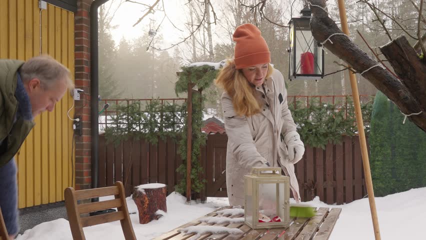 Happy winter lifestyle moment as a couple clears fresh snow from a cozy home patio. Cold weather, soft daylight, teamwork and seasonal mood create a warm family atmosphere.