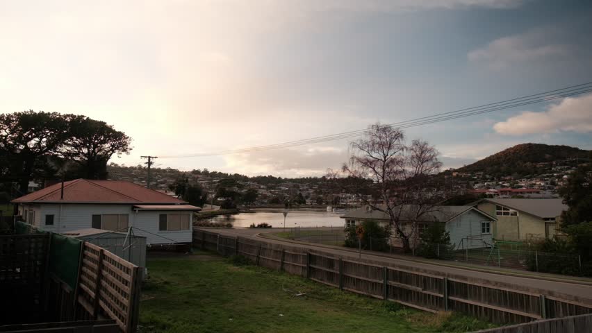Quiet suburban street scene in Hobart, Tasmania. Residential houses overlook a calm bay and hills during a peaceful sunrise. A distant person walks a dog along the water.