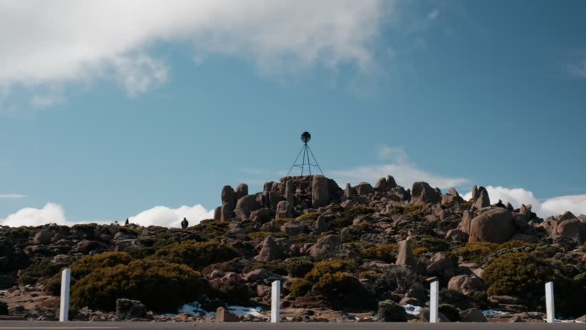 Summit of Mount Wellington (kunanyi) in Hobart, Tasmania. Rocky peak with iconic metal tripod marker and distant tourists under a blue sky.