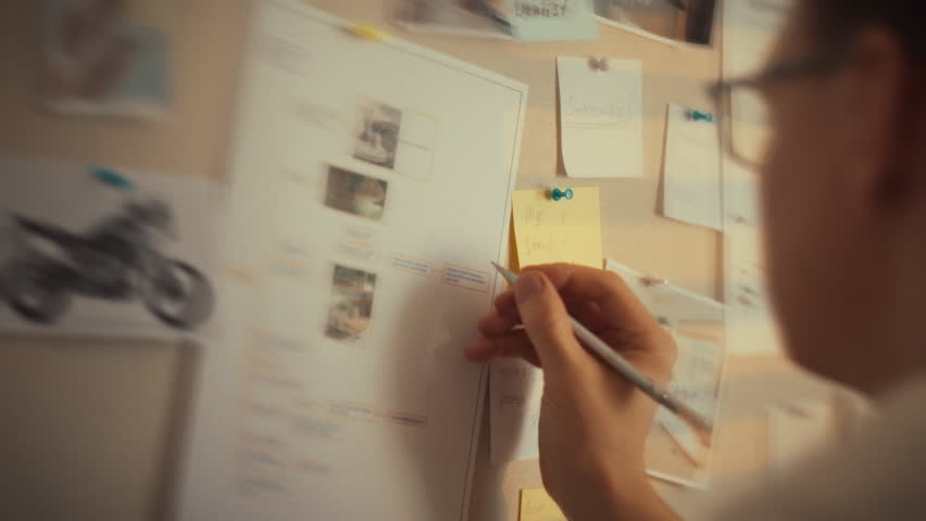A person is engaged in a brainstorming session, writing on sticky notes while surrounded by photos and notes pinned to a wall. The focus is on creativity and organization in an inspiring workspace