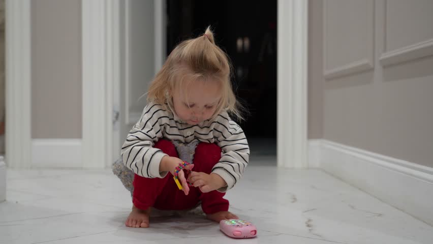Blonde child in striped top is absorbed in curious play with pink toy on serene home floor, capturing sweet moment of childhood discovery.