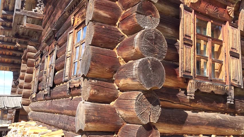 Old log house in a village with small windows featuring carved wooden frames and weathered logs. Traditional rural cottage.