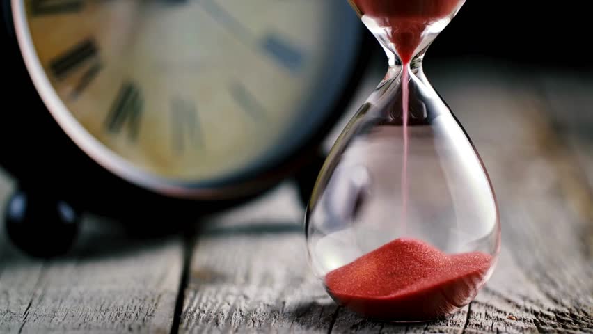 A hourglass made of glass, in the foreground with an out-of-focus clock in the background. close up of a transparent hourglass with flowing sand on blur clock background. Old classic timer.