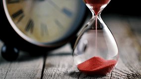 A hourglass made of glass, in the foreground with an out-of-focus clock in the background. close up of a transparent hourglass with flowing sand on blur clock background. Old classic timer. - Powered by Shutterstock - Get 15% off with code: PIKWIZARD15