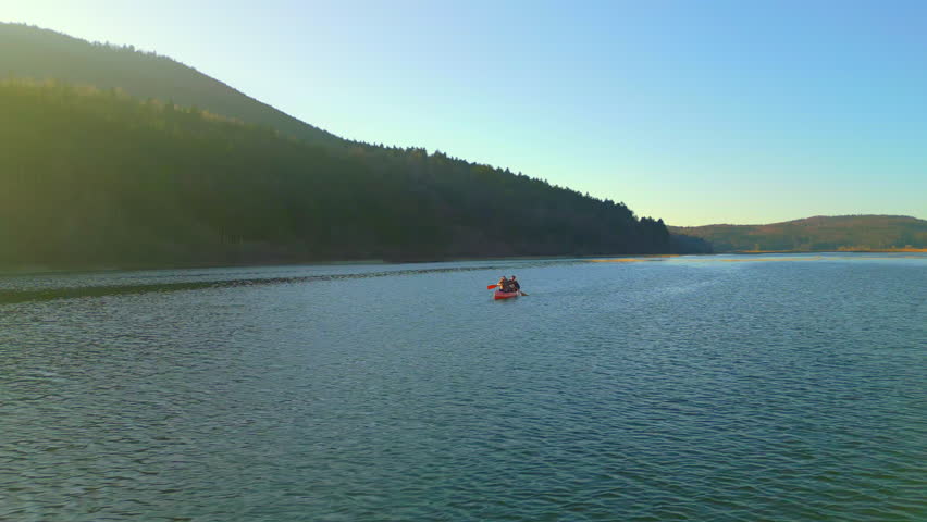 A couple paddles a red canoe on a serene lake at sunset, surrounded by forested hills. The scene captures a peaceful weekend activity, emphasizing connection, adventure, and the beauty of nature.