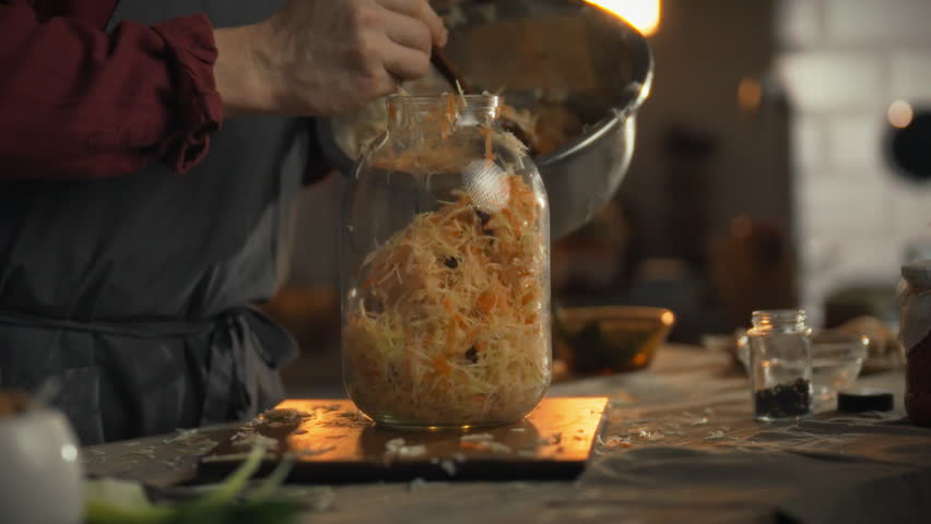 A person prepares fresh cabbage and carrots for sourdough fermentation in a warm kitchen. Ingredients are layered in a jar, showcasing traditional cooking methods and wholesome food preparation