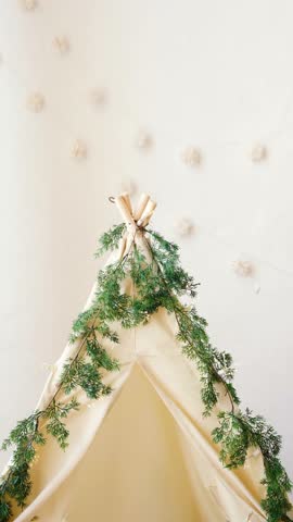 Cream teepee tent decorated with green pine garland and tiny white lights indoors.