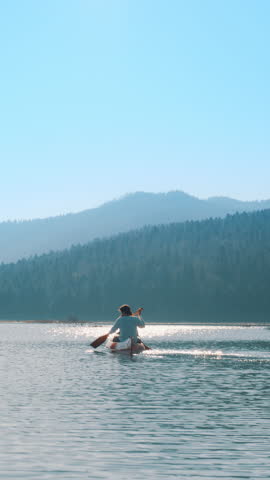 A couple paddles a canoe on a serene lake, surrounded by forested hills. The clear blue sky and calm water reflect a peaceful, active lifestyle.