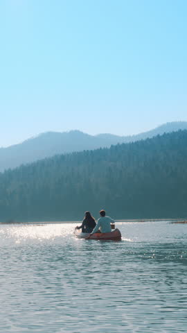 A couple paddles a canoe on a serene lake, surrounded by forested hills. The clear blue sky and calm water reflect a peaceful, active lifestyle.