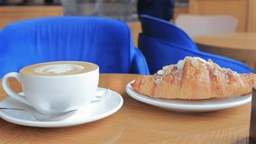 A cup of latte with foam art and an almond croissant sit on a wooden table in a modern cafe, with bright blue chairs creating a cozy and inviting atmosphere. - Powered by Shutterstock - Get 15% off with code: PIKWIZARD15