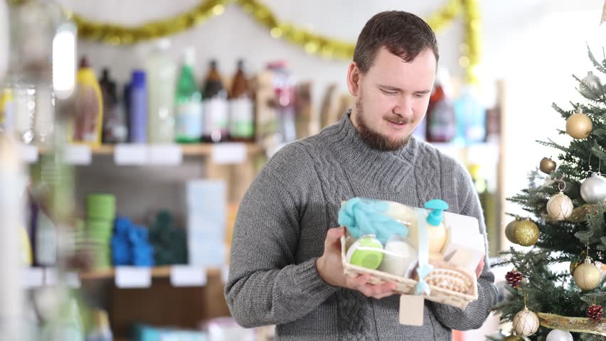 In Christmas decorated shop, man examines product and doubts, shower cosmetics present kit, study items properties in household department. Buyer examines and selects product.
