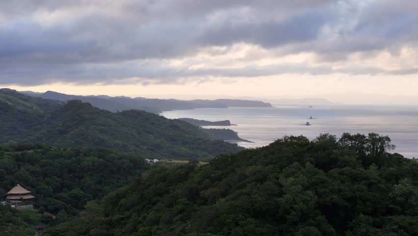 Enjoy stunning coastal landscape at dusk, featuring lush mountains and calm ocean in distance under cloudy skies.