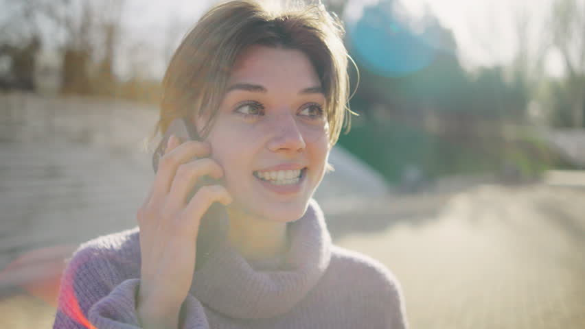 Young woman talking on the phone in a park on a sunny day while smiling happily and enjoying the moment outdoors